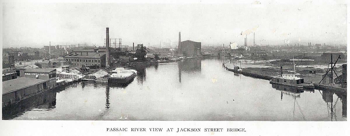 At Jackson Street Bridge
This is a view from the Jackson Street Bridge looking west. The Newark Express Company on the left is the foot of Madison Street. The Gas Tank behind the smokestack is at the corner of Market, Congress & Prospect Street. If you look closely at the background through the fog/smog you can see the skyline for that year. The right side of the photo is Harrison. The bridge you see is the railroad bridge. The photo was taken before 1912.

From "Newark - The City of Industry" Published 1912
